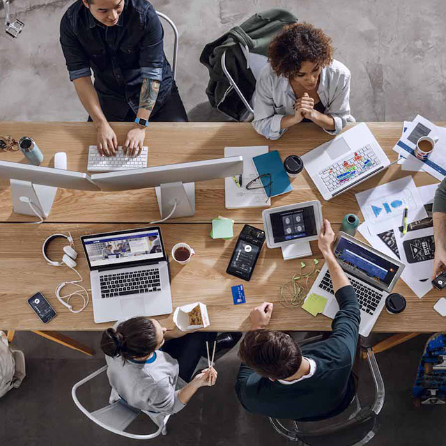 Overhead view of employees at a table with laptops, coffee mugs, notebooks, and paper spread out. 